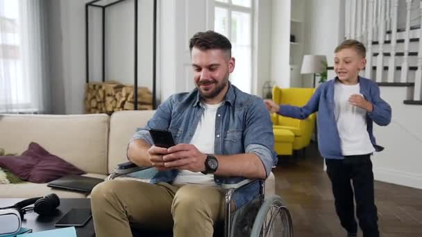 Snygg lekfull liten pojke som springer till sin godmodiga målmedvetna tillfreds skäggiga far som sitter i rullstol och surfar telefonappar — Stockvideo