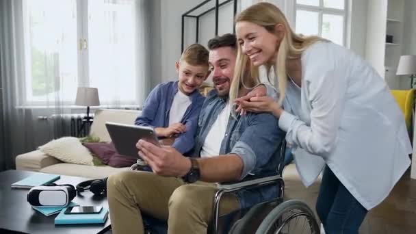 Familjebegreppet attraktiv glad leende ung mor, son och skäggig far i rullstol som har videosamtal med släktingar eller vänner från vardagsrummet — Stockvideo