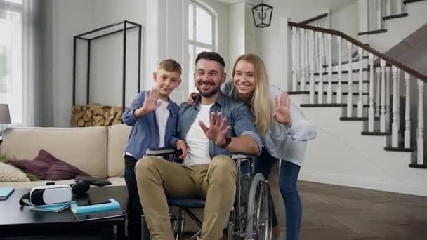 Snygg kärleksfull leende lycklig familj där unga skäggiga pappa sitter i rullstol tittar på kameran i modernt vardagsrum — Stockvideo