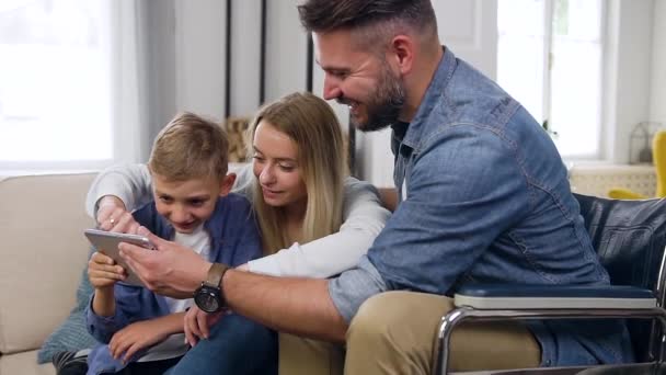 Idilio familiar donde encantador positivo feliz alegre hijo moderno, mamá y papá en silla de ruedas durante la enfermedad que disfruta de videojuegos en la tableta PC en casa — Vídeos de Stock