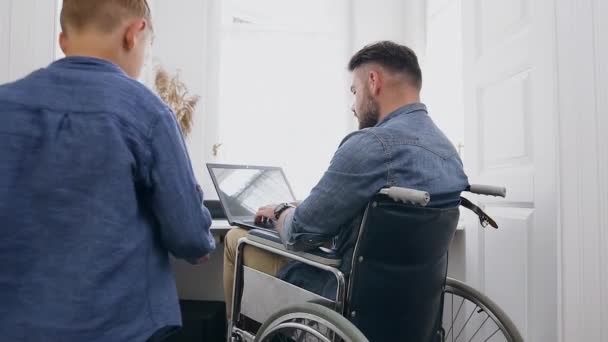 Πορτρέτο του όμορφου νεαρού γενειοφόρου άνδρα σε αναπηρική καρέκλα που μιλάει με θετικό χαμογελαστό 8χρονο γιο του κατά τη διάρκεια της μακρινής εργασίας στον υπολογιστή στο σπίτι — Αρχείο Βίντεο