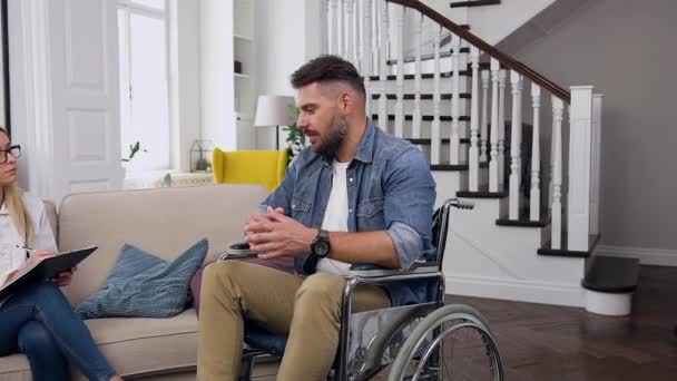 Aantrekkelijke zittende jonge bebaarde man die in een rolstoel zit en praat met attent vrouwelijk medisch assistent tijdens haar bezoek aan hem thuis — Stockvideo