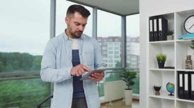 Yakışıklı, akıllı, sakallı, 30 yaşında, mühendis tasarımcı tasarım ofisinde tablet bilgisayar kullanarak gelecekteki bina hesaplamalarını özetliyor.