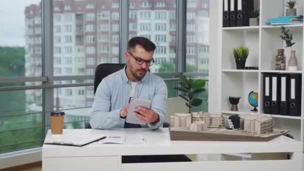 Beau concentré créatif réussi jeune concepteur barbu assis sur le lieu de travail avec modèle construit de nouveaux bâtiments et de prendre des notes dans i-pad, 4k — Video