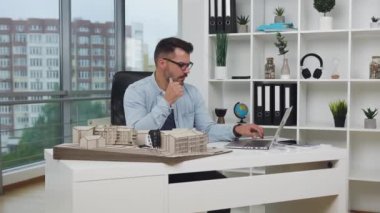 İyi görünümlü, dengeli sakallı, gözlüklü, bilgisayardan teknik ölçümleri kontrol eden, tasarım ofisindeki yeni binanın ahşap modeli, 4K