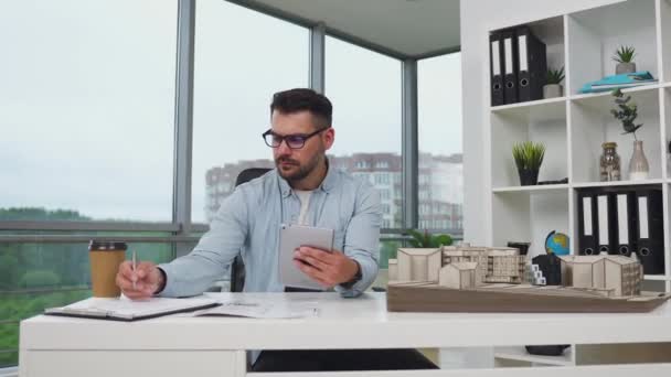 Gözlüklü, 35 yaşında, iyi görünümlü, yüksek yetenekli, sakallı bir mimar. Tablet bilgisayarından yeni binaların ölçümlerini ve tasarım ofisindeki proje belgelerini kontrol ediyor. — Stok video