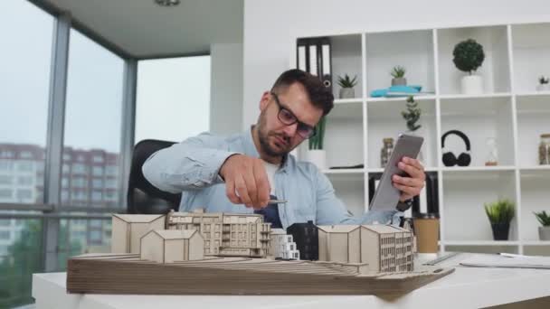 Front view of balanced attractive hardworking young bearded engineer which working with constructed model of new building checking datas from i-pad in design office,4k — Stock Video