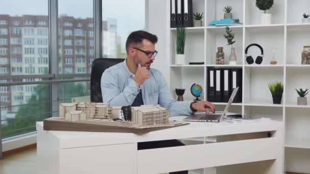 Симпатичный сбалансированный опытный бородатый архитектор в очках, проверяющий технические измерения от компьютера на деревянной модели нового здания в конструкторском бюро, 4k — стоковое видео