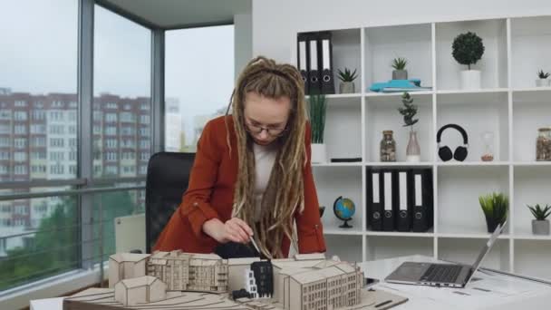Güzel görünümlü, kendinden emin, rastalı genç bayan tasarımcı modern tasarım ofisindeki gelecekteki binaların maketlerinin detaylarını kontrol ediyor. — Stok video