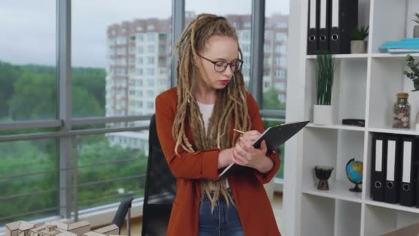 Mujer joven elegante elegante ocupada bastante concentrada con rastas haciendo notas en la carpeta sobre la construcción técnica de nuevos edificios en el estudio de diseño, 4k — Vídeo de stock