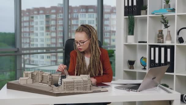 Attraente concentrato moderno esperto giovane architetto donna con dreadlocks che lavora con modello in miniatura di edifici futuri in ufficio di progettazione — Video Stock