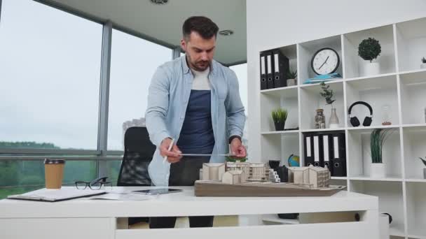 Handsome responsible hardworking young bearded engineer-designer using ruler taking measurements of constructed buildings model and writing datas into his report — Stock Video