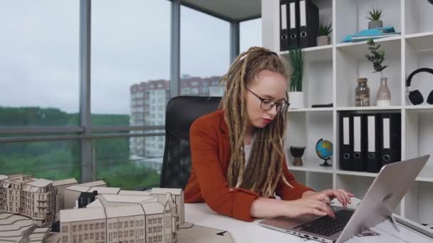 Beautiful concentrated purposeful 30-aged female architect with dreadlocks working wirh wooden construction of future buildings and special program on laptop in office — Stock Video