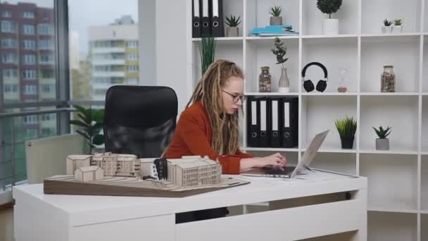 Όμορφος κομψός υψηλής ειδίκευσης γυναίκα αρχιτέκτων με αυτοπεποίθηση με dreadlocks εργάζονται σε φορητό υπολογιστή στο γραφείο σχεδιασμού και τον έλεγχο σωστές αναλογίες σε ξύλινα κτίρια μοντέλο — Αρχείο Βίντεο