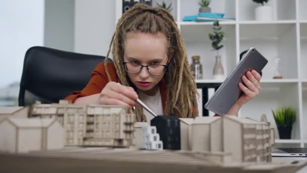 Attractive attantive modern confident young female architect with dreadlocks working with wooden model of new buildings and entering datas into tablet pc in design studio — Stock Video