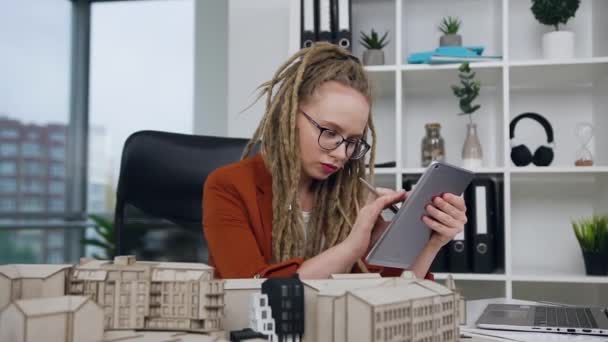 Snygg koncentrerad högkvalificerad ung kvinnlig arkitekt med dreadlocks som arbetar med i-pad och konstruerad modell av nya byggnader i designstudio — Stockvideo