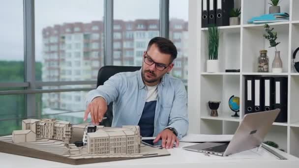 Bonito confiante criativo jovem arquiteto barbudo em óculos trabalhando no escritório de design com modelo de madeira de novos edifícios e olhando para a câmera com sorriso de sorte — Vídeo de Stock