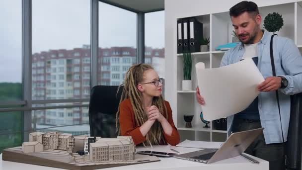 Bonito confiante e habilidoso jovem arquiteto barbudo mostrando planta para o seu charmoso chefe feminino elegante satisfeito com dreadlocks no escritório de design — Vídeo de Stock