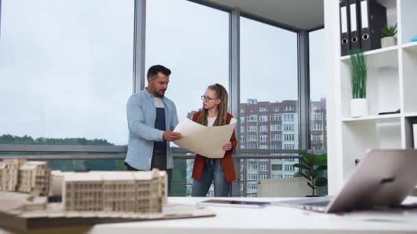 Good-looking contented creative young bearded man and woman with dreadlocks standing near the big window in design office and discussing blueprint of new buildings — Stock Video