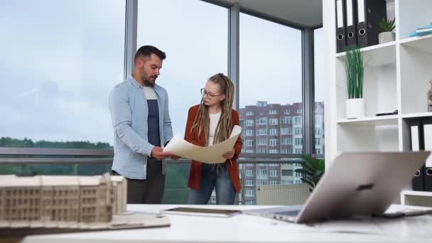 Likable positive hardworking young bearded man and woman with dreadlocks standing near design office window and discussing blueprint of new building or shopping center — Stock Video