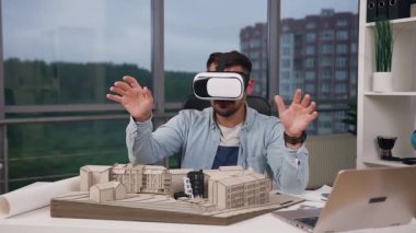 Yakışıklı tatmin edici sakallı tasarımcı iş yerinde oturmuş yeni binaların maketini yapıyor ve özel 3D gözlükler kullanarak sanal ekranda çalışıyor.