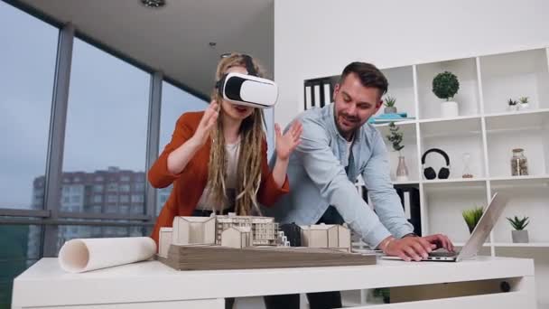 Portret van vrolijke glimlachende ervaren jonge architecten die samenwerken met houten model van toekomstige gebouwen in ontwerpbureau met behulp van informatie van laptop en augmented reality bril — Stockvideo