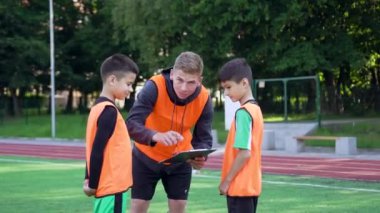 Futbol sahasındaki antrenman gününde futbol öğrencilerine maç planını açıklayan kendinden emin genç futbol koçunun ön görüntüsü, 4K