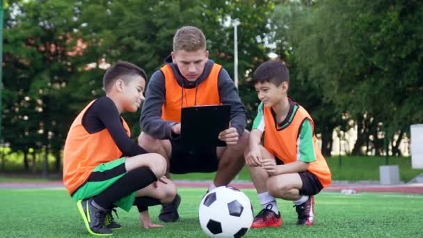 サッカー場でのトレーニング中に2人のジュニア選手にゲームのモデルを説明する本格的な経験豊富な若いサッカーコーチのフロントビュー, 4k — ストック動画