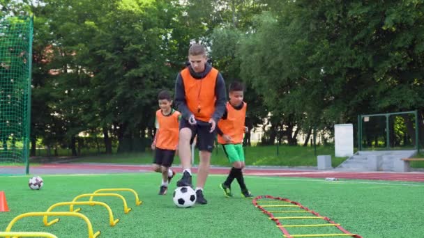 Koncepcja piłki nożnej, gdzie dwóch ufnych nastolatków w pomarańczowych kamizelkach ćwiczy piłkę nożną pod dowództwem młodego, doświadczonego trenera na stadionie. — Wideo stockowe