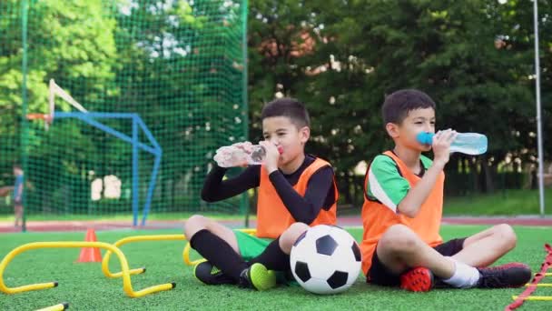 Vista frontal de bonitos jogadores de futebol adolescente cansados que se sentam na cobertura especial verde no campo esportivo e bebem água doce durante o treinamento ao ar livre — Vídeo de Stock