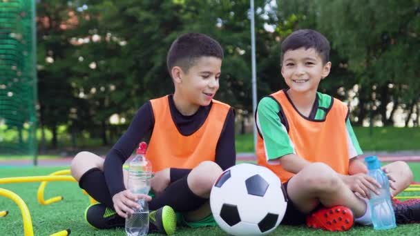 Gustable sonriente satisfecho jugadores de fútbol junior sentados en el terreno de césped verde y beber agua de la botella después del entrenamiento de fútbol, 4k — Vídeos de Stock