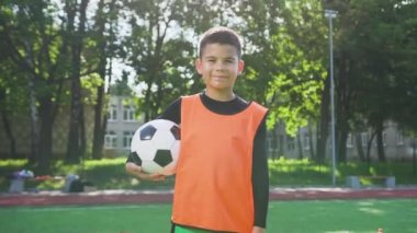 Çekici, mutlu, turuncu yelekli genç futbolcu elinde topla dışarıda spor arka planında kameraya bakıyor.