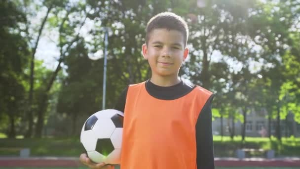 Goedhartige vrolijke sportieve tiener jongen in voetbal uniform houden bal in zijn handen en poseren op camera op de buitenlucht sportveld, 4k — Stockvideo