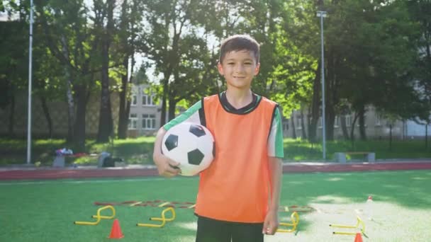 屋外のサッカー場の背景にボールとカメラにポーズオレンジベストで見栄えの良い十代のサッカー選手のフロントビュー — ストック動画