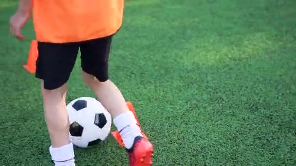 Крупный план мальчишеских ног, которые делают беговые упражнения с мячом вокруг стойки на тренировочном футбольном поле на открытом футбольном поле — стоковое видео