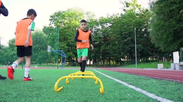 Esportivo intencional adolescente meninos em uniformes de futebol fazendo exercícios de corrida com a superação de obstáculos sob o comando de seu treinador experiente responsável durante o treinamento no estádio — Vídeo de Stock