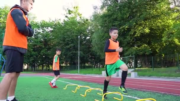 Retrato de jogadores de futebol 13s persistentes concentrados que fazendo exercícios de corrida com obstáculos no estádio com relva artificial sob o comando de treinador jovem experiente — Vídeo de Stock