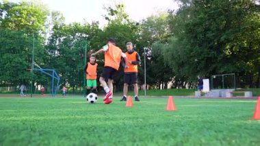 İyi görünümlü, yetenekli genç bir futbol hocası genç öğrencilerinin açık hava spor sahasında antrenman yaparken engelleri kullanarak topa nasıl koştuğunu izliyor.