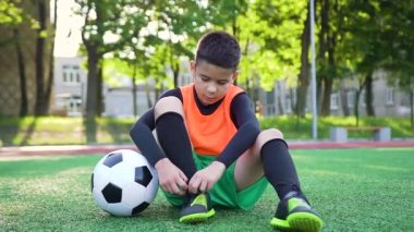 Yeşil çimlerde oturan ve ayakkabı bağcığını bağlayan futbol formalı yakışıklı, kendine güvenen genç çocuk.