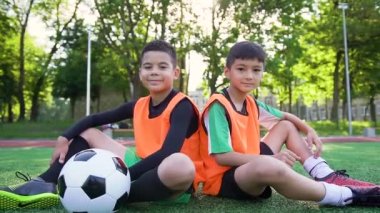 İyi huylu, güler yüzlü, turuncu yelekli iki futbolcu suni sahada oturuyor ve baş parmaklarıyla kameraya poz veriyor.