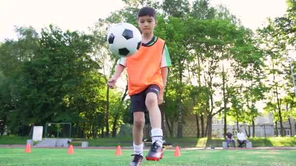 Портрет симпатичного уверенного подростка в футбольной форме с оранжевым жилетом, который набивает мяч ногами дрессировкой на открытом спортивном поле в летний день — стоковое видео