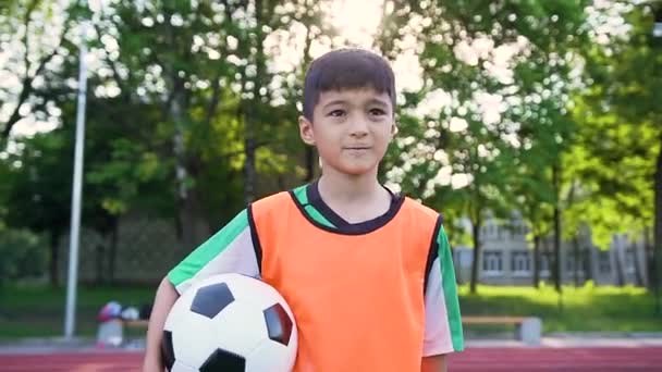 训练开始前，穿着橙色背心站在室外运动场上拿球的英俊而饱满的少年足球运动员 — 图库视频影像