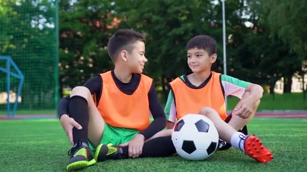 Atraente sorrindo esportivo dois jogadores de futebol sentados na cobertura verde artificial do campo de esporte ao ar livre e dando alta cinco uns aos outros — Vídeo de Stock