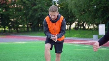 Yüksek yetenekli genç futbol antrenörü, yetenekli öğrencileri açık hava spor sahasında hız egzersizleri yaparken zamanı kontrol ediyor.