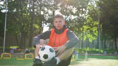 Yakışıklı, kendine güvenen genç futbol koçu açık hava spor sahasının suni sahasında oturuyor ve kameraya bakıyor.