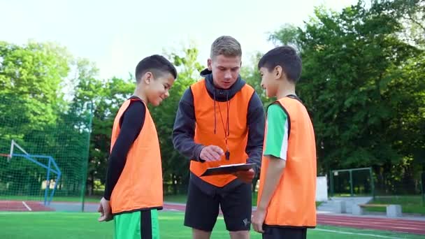 Close up de concentrado bem parecido jovem treinador de futebol em uniforme que explicando o modelo de jogo para seus alunos adolescentes sorridentes no campo de futebol — Vídeo de Stock