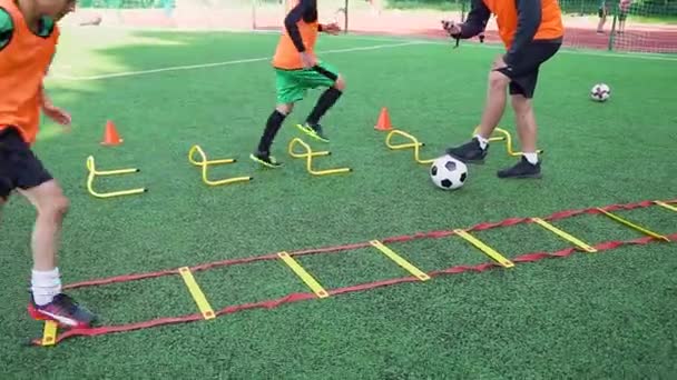 Niños adolescentes enérgicos persistentes en ropa de fútbol haciendo ejercicios de carrera con obstáculos bajo el mando de su instructor altamente calificado en el campo de deporte — Vídeo de stock