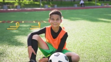 Spor sahasının suni sahasında oturup kameraya güzel bir gülümsemeyle bakan yakışıklı, esmer, genç futbolcu.