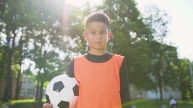 Turuncu yelek giymiş, elinde top tutan ve güneşli bir günde açık hava spor sahasındaki kameraya bakan 13 yaşında hoş bir futbolcu.