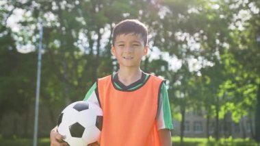 Şehir parkındaki stadyumdaki futbol antrenmanında topla kameraya poz veren futbol takımındaki kendinden emin genç çocuğun ön görüntüsü.